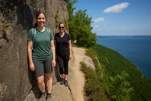 Two women hiking along the Precipice Trail Acadia National Park on a narrow granite ledge overlooking Frenchman Bay under bright summer skies.
